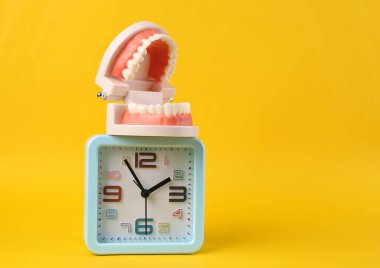 Sarı arkaplanda alarm saati olan bir insan çenesinin plastik modeli