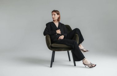 Stüdyodaki koltukta oturan siyah takım elbiseli şık bir kadının moda çekimi. Vogue, Yaşam Tarzı