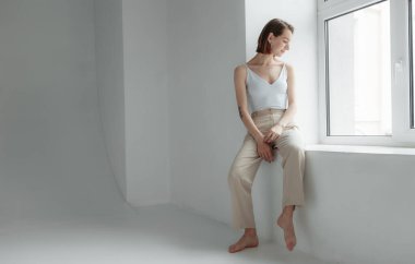 Beyaz bir odada, ışık penceresinin yanında oturan şık bir kadın.