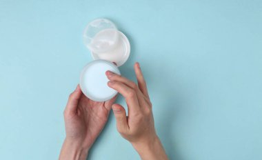 Kadın elleri mavi arka planda bir kavanoz krema tutuyor.