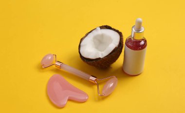 Bir şişe yüz serumu, masaj rulosu ve sarı arka planda hindistan cevizi. Doğal kozmetik konsepti