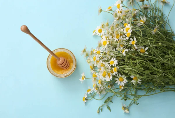 Mavi arka planda kaşıkla bir kavanoz bal ve bir buket papatya
