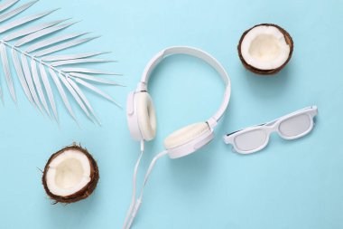 Mavi arka planda beyaz güneş gözlüğü, kulaklık, hindistan cevizi ve palmiye yaprakları. Minimum seyahat, tropikal yerleşim. Üst görünüm