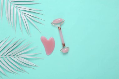 Beyaz palmiye yapraklı mavi arka planda yüz silindiri ve gua sha taş masaj aleti. Mavi arka plan. Üst görünüm