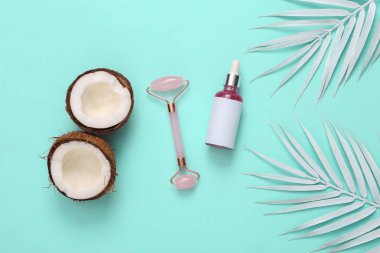 Face serum bottle and massage roller with white palm leaves and coconuts on a turquoise background. Natural cosmetics concept