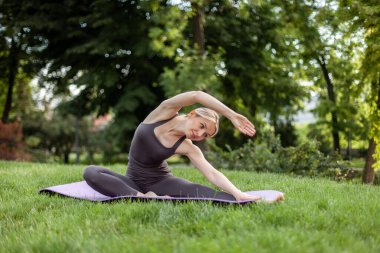 Genç ve çekici sarışın kadın parkta minderde otururken yoga pozu veriyor.