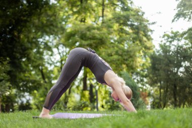 Genç ve çekici sarışın kadın parkta minderde otururken yoga pozu veriyor.