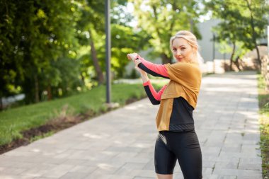 Spor kıyafetli genç bir kadın parkta esneme egzersizi yapıyor.
