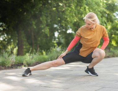 Spor kıyafetli genç bir kadın parkta esneme egzersizi yapıyor.