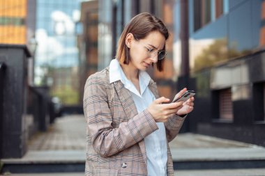 Ceketli ve gözlüklü genç, modern iş kadını şehirdeki bir akıllı telefonun ekranına bakıyor.