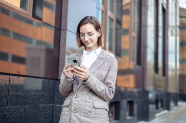 Ceketli ve gözlüklü genç, modern iş kadını şehirdeki bir akıllı telefonun ekranına bakıyor.
