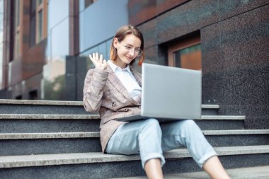 Genç iş kadını merdivenlerde dizüstü bilgisayarla otururken video görüşmesi yapıyor. Video konferansı