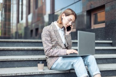 Genç modern iş kadınları müdürü şehirdeki merdivenlerde otururken dizüstü bilgisayar kullanıyor ve telefonla konuşuyor.