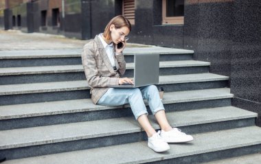 Genç modern iş kadınları müdürü şehirdeki merdivenlerde otururken dizüstü bilgisayar kullanıyor ve telefonla konuşuyor.