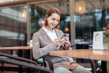 Genç bir kadın açık bir kafedeki masada otururken telefon kullanıyor.