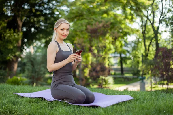 Sevimli sarışın kadın parkta minderde otururken akıllı telefon kullanıyor.