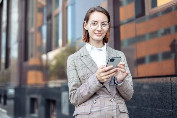 Şehirde ceketli ve gözlüklü genç, modern iş kadını akıllı telefon tutuyor.