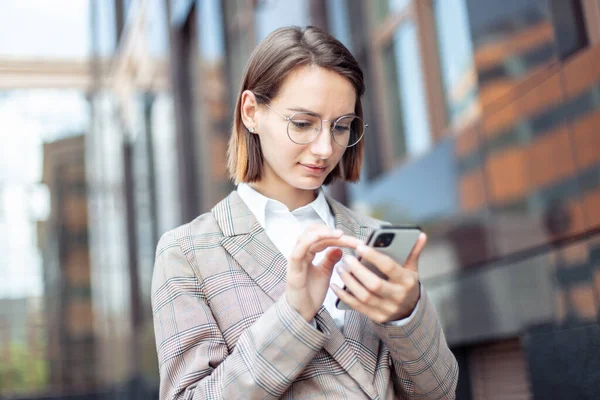 Genç, modern iş kadını iş binasının arka planında akıllı telefon kullanıyor.