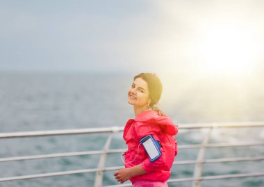 Spor giyimli kadın koşucu kamera önünde poz veriyor ve plajda kulaklıkla müzik dinliyor.