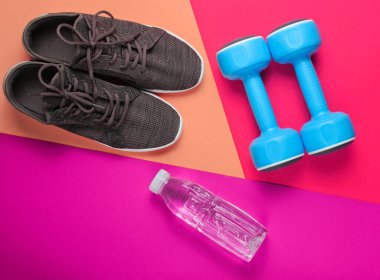 Minimalist fitness konsepti. Dambıllar, spor ayakkabılar, renkli arka planda bir şişe su. Üst görünüm