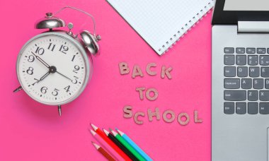 Okul zamanı minimalist konsepti. Laptop, renkli kalemler, not defteri, pembe zemin üzerindeki çalar saat. Slogan: Tahta harflerle okula dönüş, üst manzara