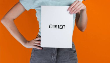 Woman holding white empty sheet of paper for copy space on orange background. Crop photo, studio shot