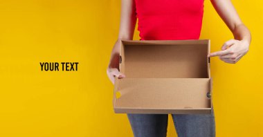 A young woman holds an empty cardboard box and points her finger inside on a yellow background. Crop photo, studio shot. Copy space