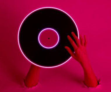 Female hands holding vinyl record with neon glow circle through the torn holes. Pink light. Retro wave. 80s