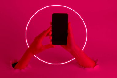 Female hands holding smartphone through the torn holes with neon glow circle. Pink light. Creative art