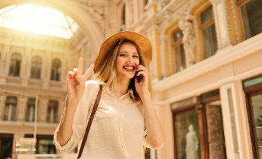 Telefondan konuşan keçeli şapkalı neşeli sarışın kadın ve eski şehir mimarisinin arka planındaki kameraya bak.