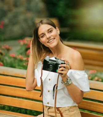 Parkta bankta otururken elinde retro kamera tutan yaz kıyafetleri içinde mutlu güzel bir kadın.