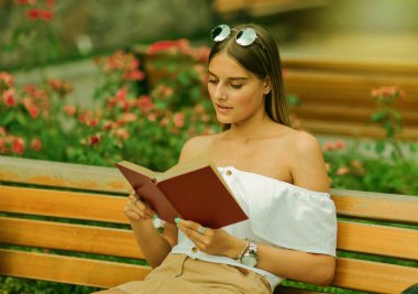 Genç sarışın bir kadın parkta bankta otururken heyecanla kitap okuyor.