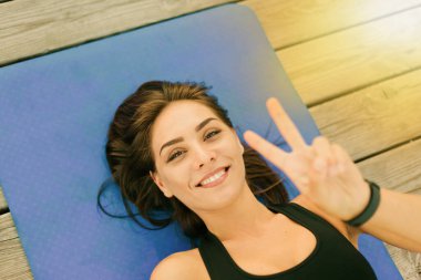 Genç, neşeli spor kadın mindere uzanıyor ve V sembolü gösteriyor. Üst görünüm portresi