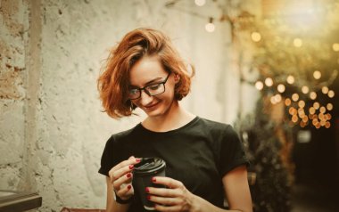 Cheerful hipster girl in glasses with short haircuts holding coffee cup against the background of street garlands outdoor