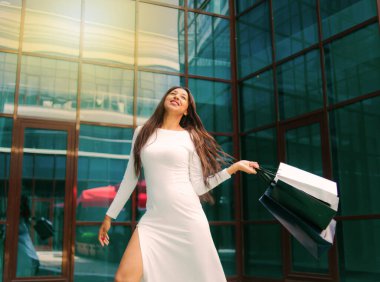 Shopping concept. Beauty afro woman in white beautiful dress holding many paper shopping bags on background of business building with blue windows