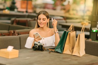 İnternetten alışveriş. Neşeli kadın alışveriş poşetleriyle masada oturuyor ve elinde akıllı telefonu ve açık bir kafede banka kartını tutuyor.