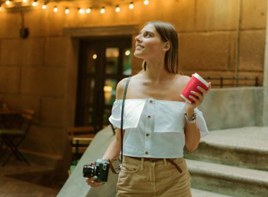 Şık giyinmiş güzel sarışın bir kadın. Elinde retro kamera ve kahve fincanı var. Dışarıda, çelenklerin arkasında.