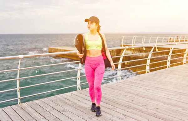 Spor giyimli genç bir kadın ve kumsalda yürürken omzunda bir çanta.