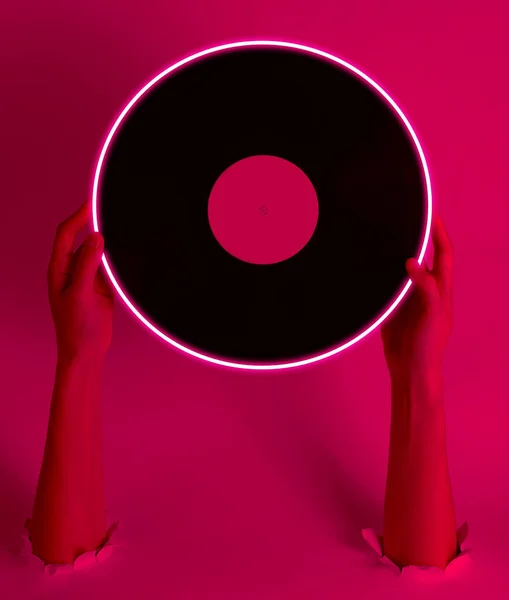 Female hands holding vinyl record with neon glow circle through the torn holes. Pink light. Retro wave. 80s