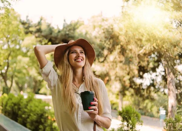 Cheerful blond woman in casual style clothes and felt hat drinking coffee on the go and walking in the city