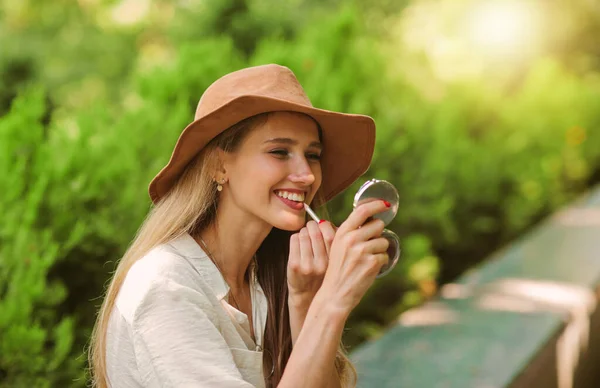 평상복을 모자를 기분좋은 금발의 여자는 립라이너로 화장을 거울을 — 스톡 사진