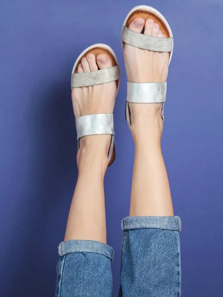 Raised Female Legs Blue Jeans Trendy Leather Sandals Blue Background — Stockfoto