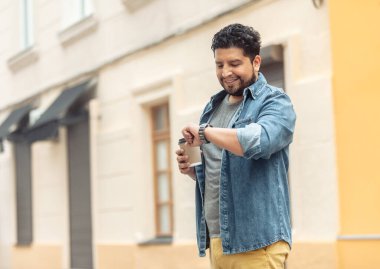 Yakışıklı, gülümseyen İspanyol adam kahve içiyor ve şehirde saatine bakıyor.