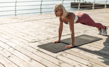 Spor kıyafetli genç sarışın kadın plajda şınav çekiyor.