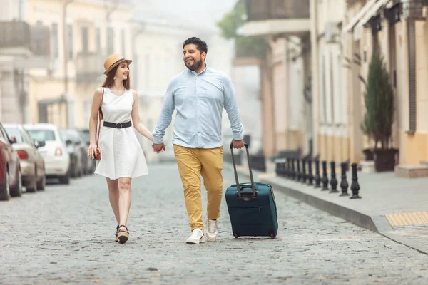 Joint, romantic trip. Loving international couple of tourists with travel luggage are walking along a European street. Spend time together.