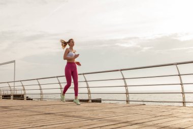 Spor kıyafetli genç bir kadın sabahın erken saatlerinde sahilde koşuyor. Sağlıklı yaşam tarzı, kardiyo çalışması