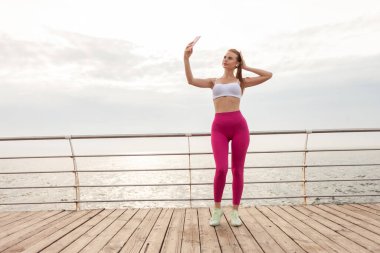 Spor kıyafetlerindeki güzel kızıl saçlı fitness modeli sahildeki bir akıllı telefondan selfie çekiyor.
