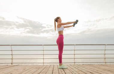 Spor kıyafetlerindeki kızıl saçlı fitness mankeni plajda halterle çalışıyor. Sağlıklı yaşam tarzı