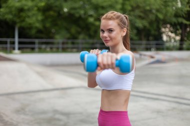 Kızıl saçlı, formalı, spor kıyafetli bir kadın. Dışarıda halterlerle çalışıyor. Sağlıklı yaşam tarzı kavramı