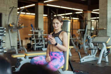 Formda bir kadın spor salonundaki bankta otururken akıllı telefon kullanır.